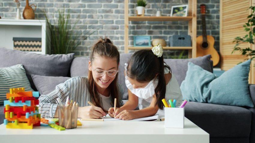 Happy little girl is drawing at home with caring mother using pencils having fun in modern apartment. Creative leisure activity and childhood concept.